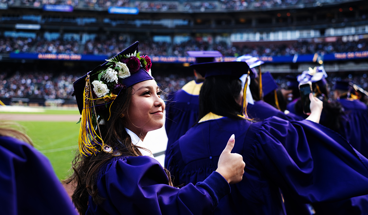Student dressed in regalia in a ballpark gives the thumbs up 